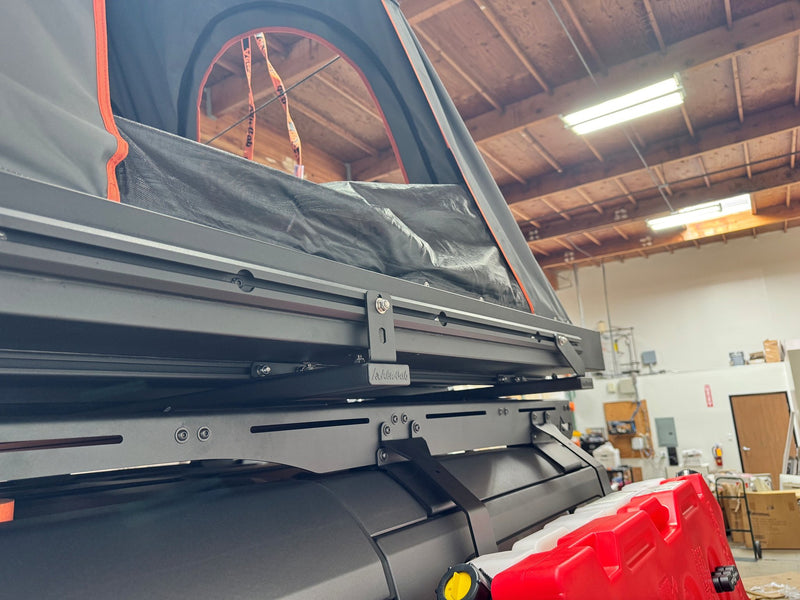 Side view of the Alu-Cab Gen 3-R rooftop tent partly opened, showing the Alu-Cab branded mounting hardware, the rack rails it sits on and the mattress stowed inside the shell — photo 4