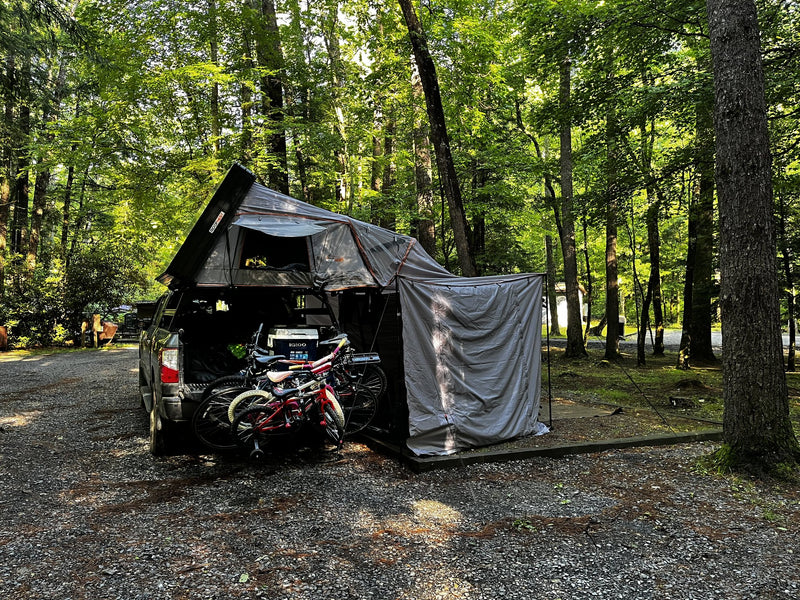 Forest campsite with the tent open on the truck and the Awnex awning room fully enclosed with all four walls zipped on, bikes and a cooler staged in the truck bed alongside — photo 4