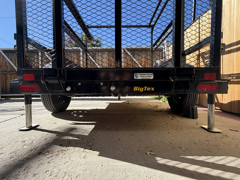 Low rear view beneath the trailer showing both tires, the axle, the rear stabilizing jacks extended to the ground and the Big Tex rear badge — photo 12
