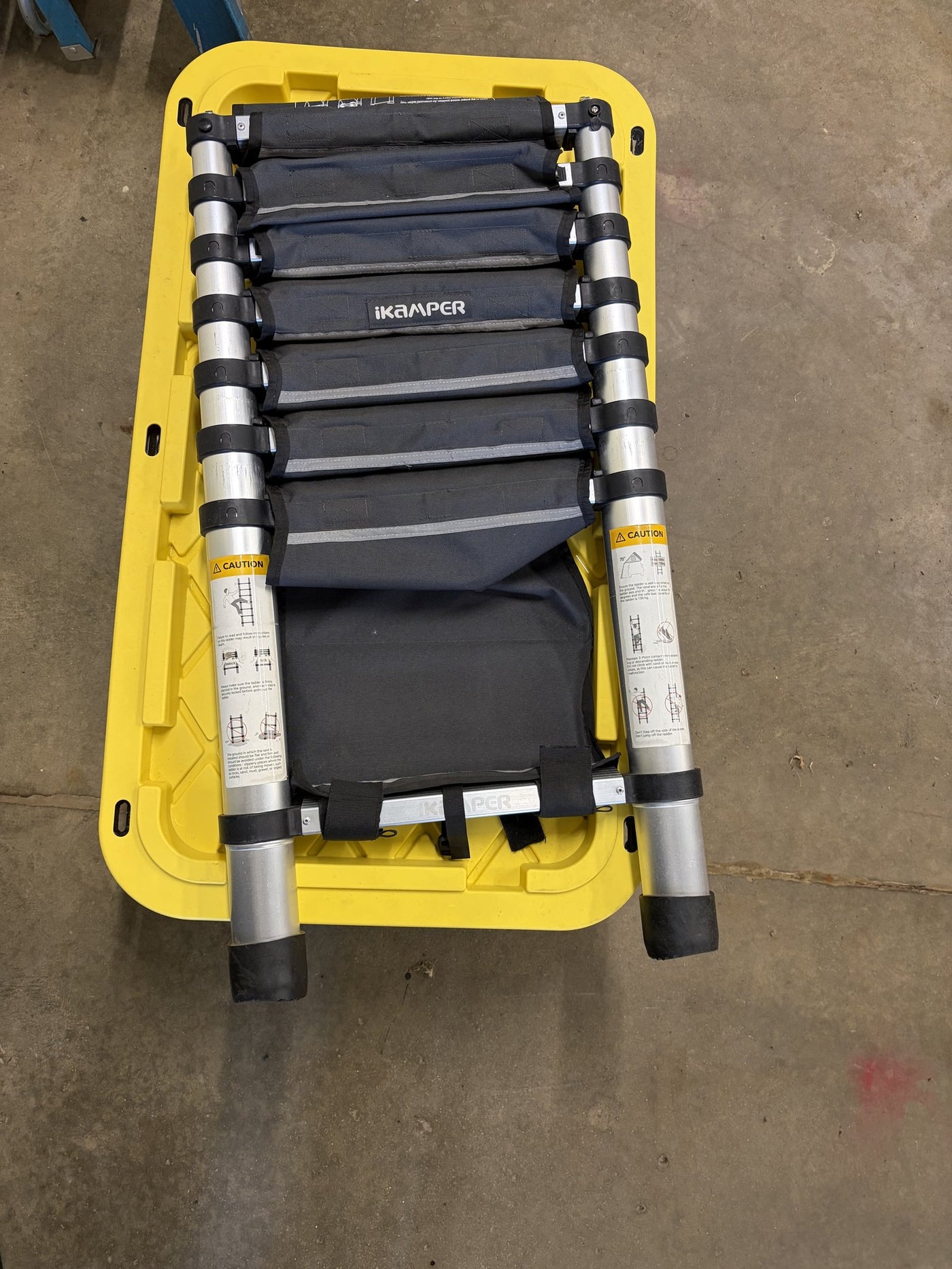 The iKamper telescoping aluminium ladder with the padded fabric steps fitted over the rungs, folded into a storage tote with its caution labels visible — photo 10