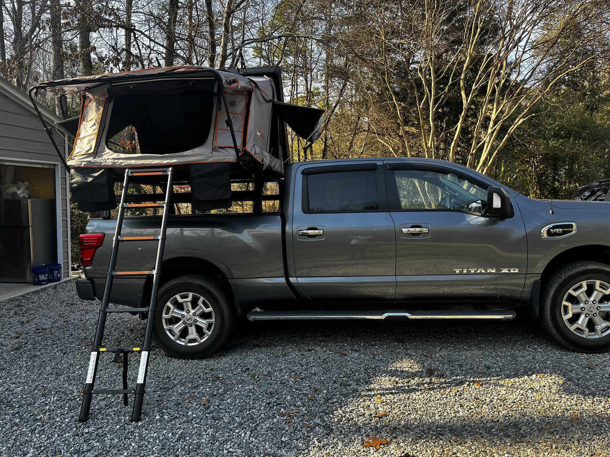 Roofnest Condor Overland 2XL rooftop tent opened on a bed rack over a grey Nissan Titan XD in a gravel driveway, grey and orange fold-out tent body with large mesh door panels and the telescoping ladder extended to the ground — photo 1