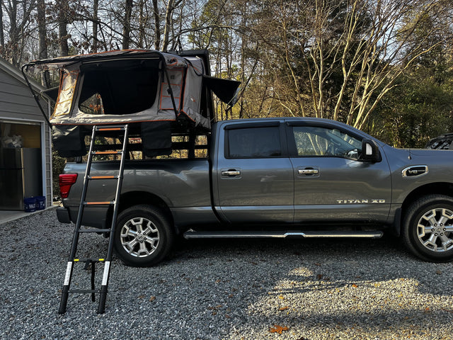 Roofnest Condor Overland 2XL rooftop tent opened on a bed rack over a grey Nissan Titan XD in a gravel driveway, grey and orange fold-out tent body with large mesh door panels and the telescoping ladder extended to the ground