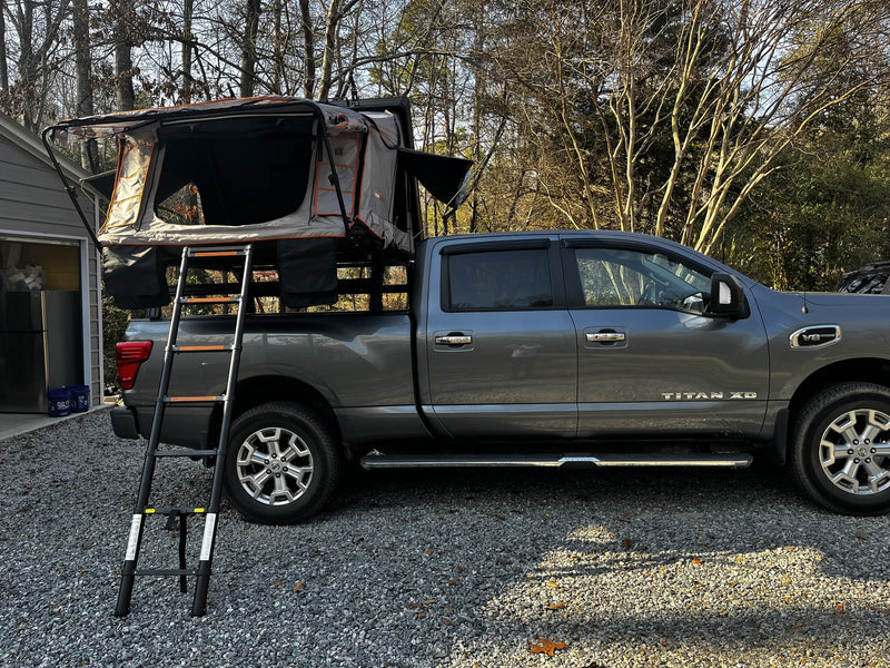 Roofnest Condor Overland 2XL rooftop tent opened on a bed rack over a grey Nissan Titan XD in a gravel driveway, grey and orange fold-out tent body with large mesh door panels and the telescoping ladder extended to the ground — photo 1