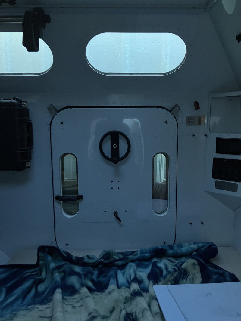 Interior of the 2023 Mammoth Overland ELE looking at the rear hatch: white composite cabin walls, oval porthole windows, a latching rear door with a round handle, a bear spray bracket, a hard case, and bedding on the sleeping platform — photo 6