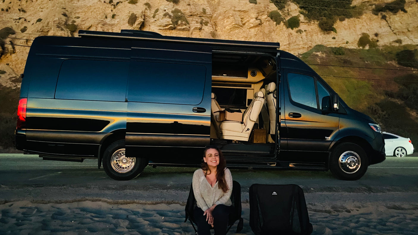 Smiling woman by a luxury black RV rental with open door revealing interior, parked by the beach. Perfect outdoor living.