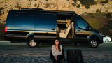 Smiling woman by a luxury black RV rental with open door revealing interior, parked by the beach. Perfect outdoor living.