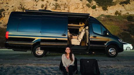 Smiling woman by a luxury black RV rental with open door revealing interior, parked by the beach. Perfect outdoor living.