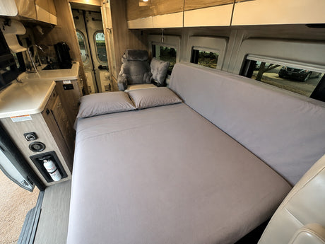Spacious RV rental interior with a neatly made bed, kitchenette, and passenger seat. Fire extinguisher visible for safety.