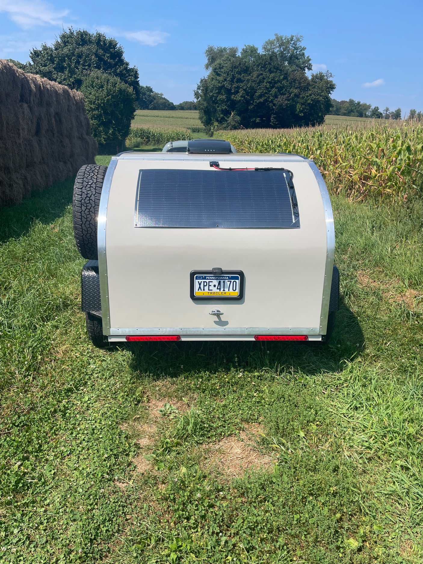 Teardrop RV rental with solar panel & spare tire on grass. Rural field, hay bales, and corn await your outdoor adventure.