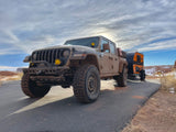 A mud-splattered Jeep Gladiator tows an orange RV rental on an asphalt road, set against a desert mountain backdrop.