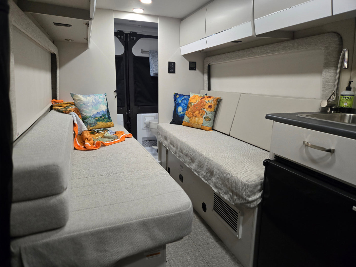 Cozy RV rental interior: two facing benches with patterned upholstery, art pillows, compact kitchen, sink, and storage.