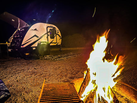 An RV rental camper illuminates a dark campsite beside a crackling campfire, offering warmth on a cool night.