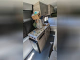 This RV rental interior shows a kitchenette with a floral counter, fridge, and overhead cabinets, alongside a patterned bench seat.