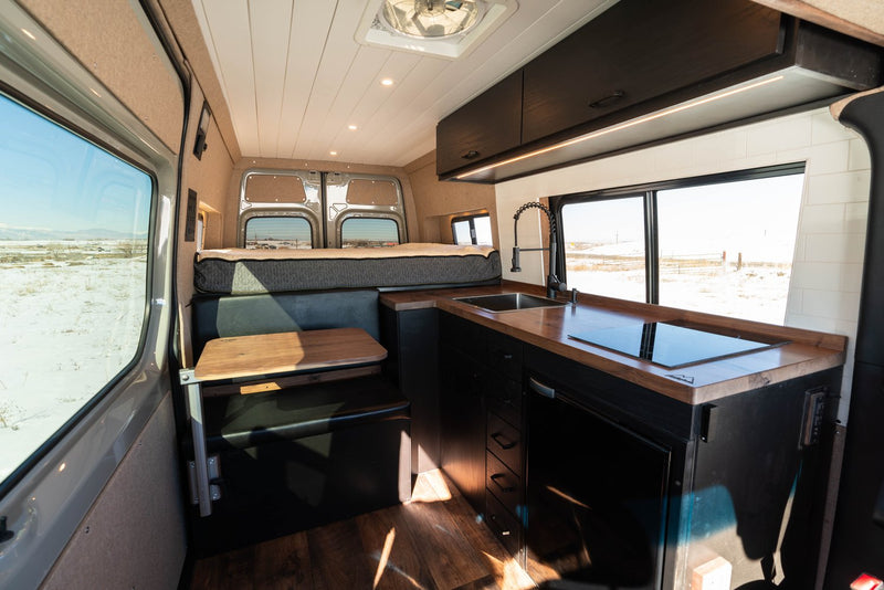  and a scenic snowy mountain view from the window. Perfect for your next adventure.|This RV rental's versatile interior features a lifted bench seat revealing ample storage and a compact fold-down table.|View inside an RV rental camper van: modern kitchen with dark cabinets — photo 10