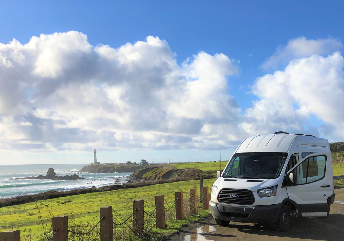 White RV rental by the coast with a distant lighthouse, lush green fields, and a blue sky with clouds.
