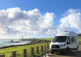 White RV rental by the coast with a distant lighthouse, lush green fields, and a blue sky with clouds.