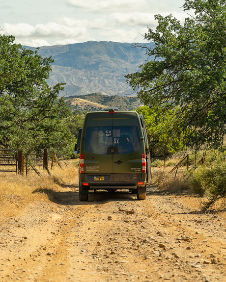 Green Suha Van RV rental on a dirt road, leading towards mountains. Perfect for off-grid adventures & nature exploration.