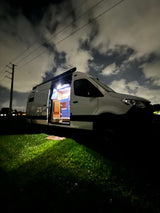 Night scene: a white rv rental camper van on grass, side door open to reveal a brightly lit, cozy interior.