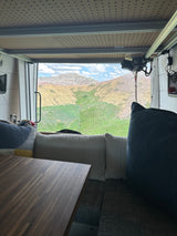 RV rental interior: a cozy dinette with a wooden table and soft cushions, offering panoramic views of colorful mountains and a green valley.