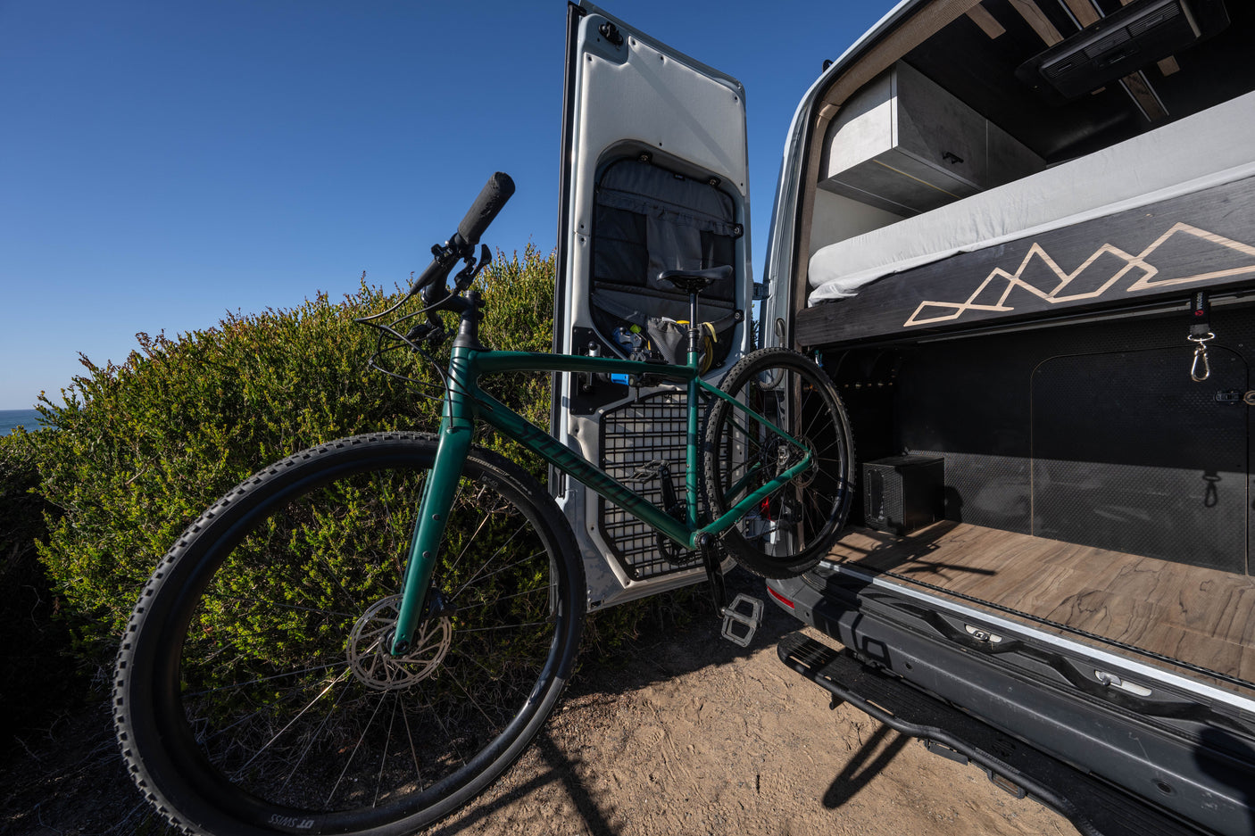 Green gravel bike positioned by a custom RV rental's open back, revealing its adventure-ready interior and bed.