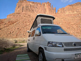 An RV rental pop-top camper van faces towering red rock cliffs, parked on a dirt path with a striped mat. Ideal for desert adventures.