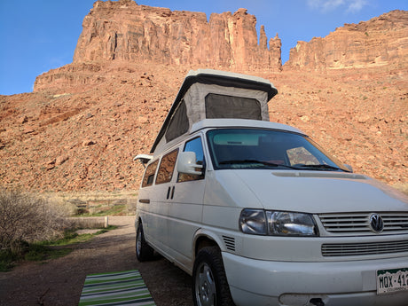 An RV rental pop-top camper van faces towering red rock cliffs, parked on a dirt path with a striped mat. Ideal for desert adventures.