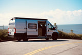 A silver RV rental camper van is parked on a coastal road with the ocean in the background, side door open.