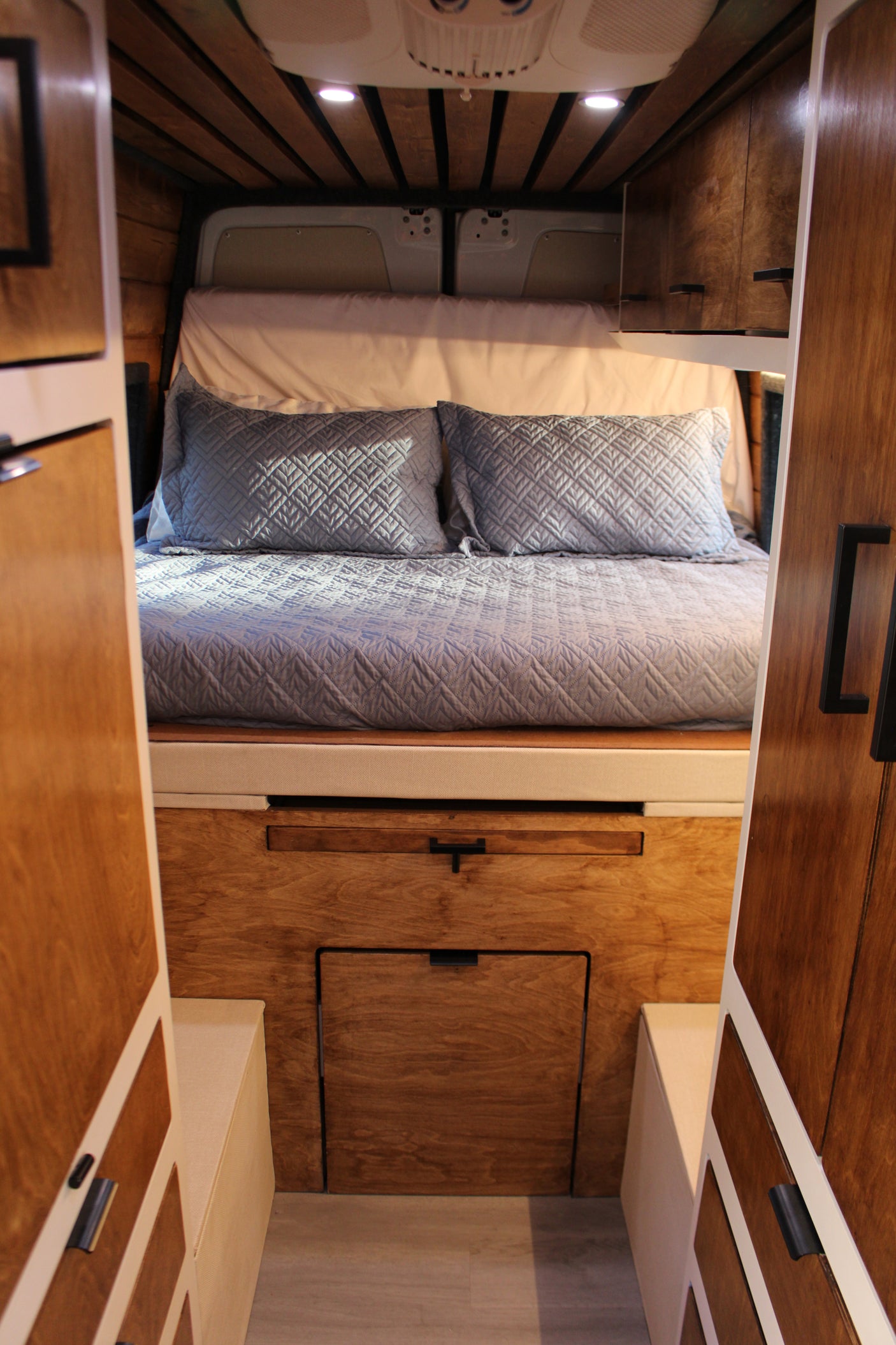 Plush bed in a cozy RV rental, featuring warm wood cabinetry, overhead lighting, and smart storage.