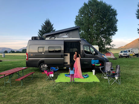 Family preparing camp with their **RV rental**, a gray camper van with pop-top, at a grassy campsite.