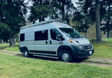 Silver Ram ProMaster RV rental, with an awning, parked on a gravel path by tall evergreen trees.