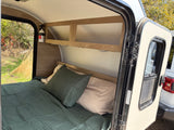 Cozy RV rental interior with a comfortable bed, overhead shelf, and a view of nature outside the open door. Ideal for camping.