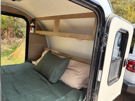 Cozy RV rental interior with a comfortable bed, overhead shelf, and a view of nature outside the open door. Ideal for camping.