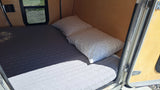 Inside a compact rv rental, a bed with a quilted mattress and two pillows. Sunlight creates shadows on the sleeping area.