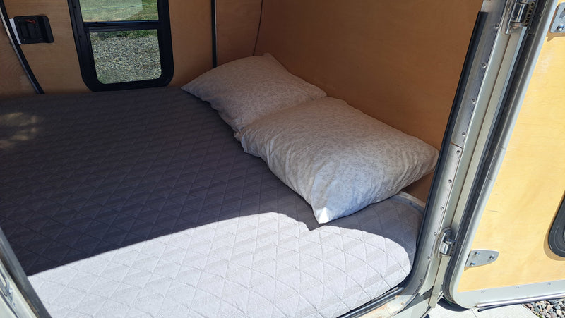 Inside a compact rv rental, a bed with a quilted mattress and two pillows. Sunlight creates shadows on the sleeping area. — photo 10