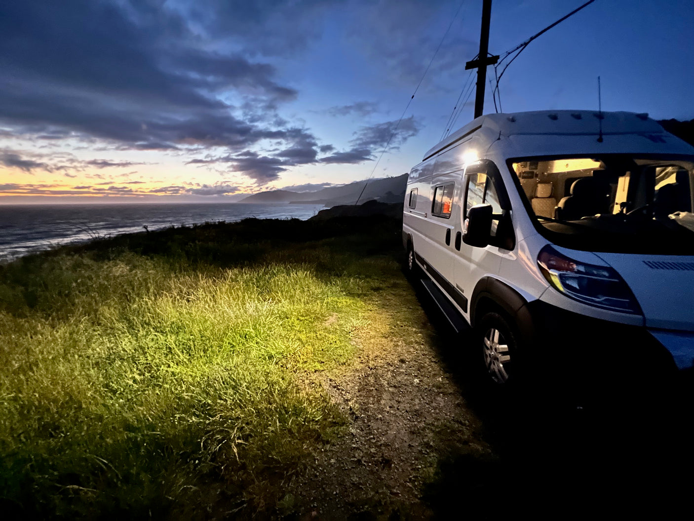 RV rental parked at dusk by the ocean, its light illuminating the grassy area. Scenic sunset over the water.