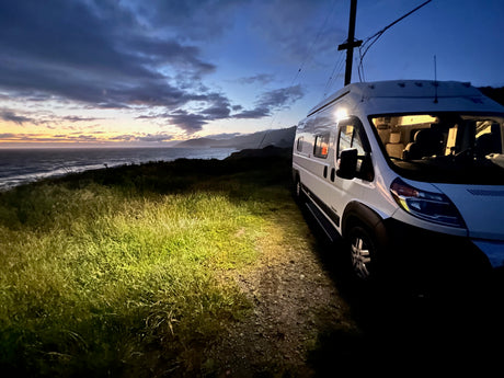RV rental parked at dusk by the ocean, its light illuminating the grassy area. Scenic sunset over the water.