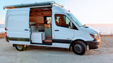 White Mercedes Sprinter campervan, ideal for an RV rental, parked on a coast with open door revealing its custom interior.