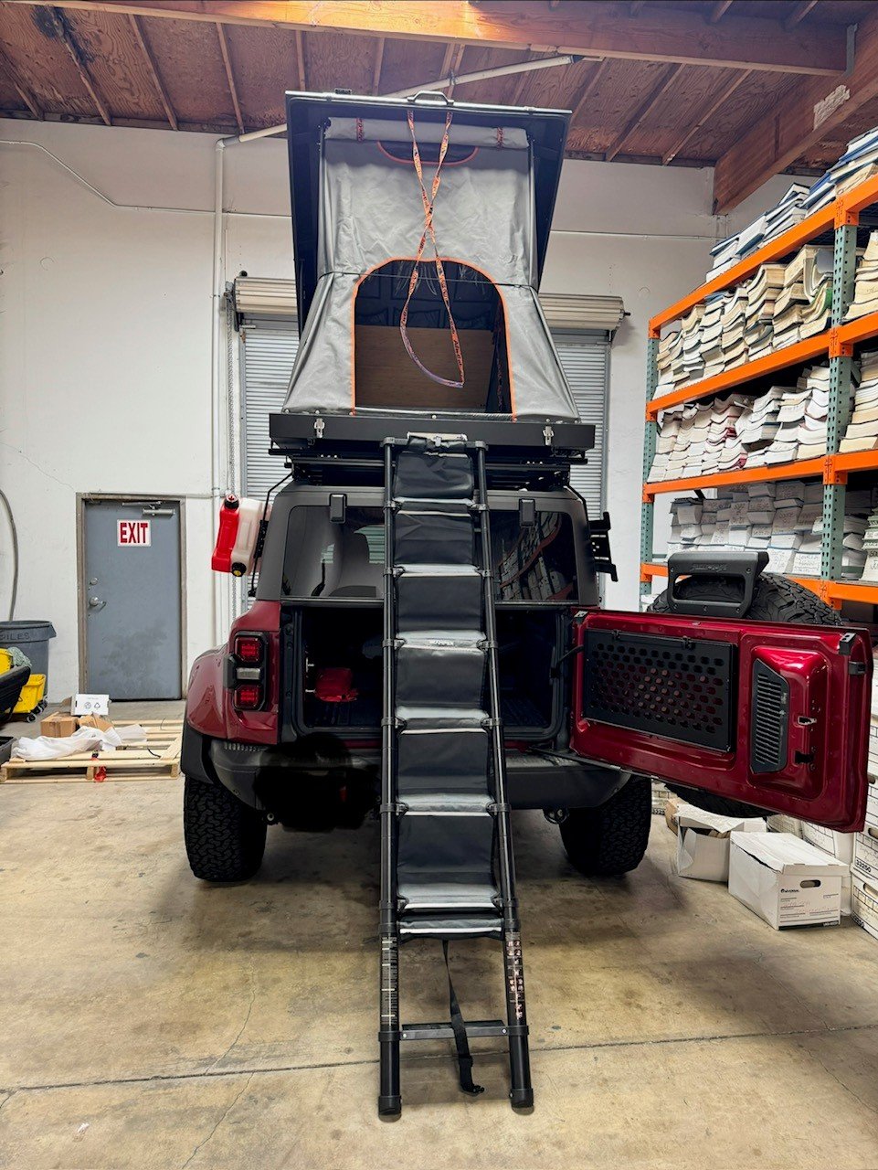 Rear view of the Alu-Cab Gen 3-R rooftop tent opened on a red Land Rover Defender with the telescoping ladder extended to the ground, showing the tent's entry, interior bamboo panel and the vehicle's open rear door — photo 2