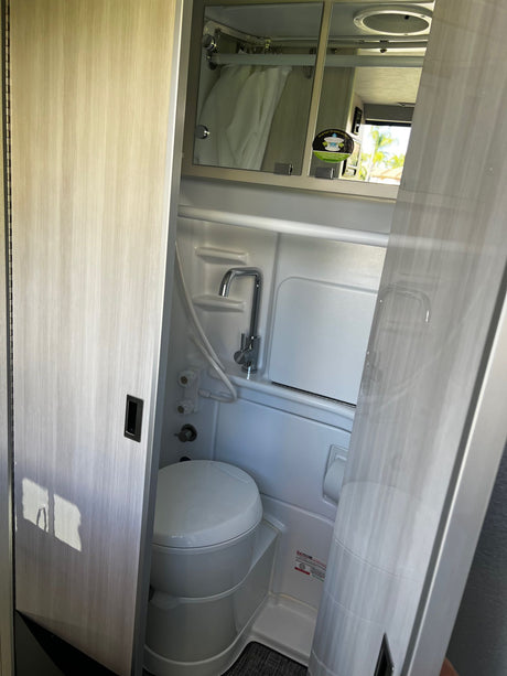 Compact wet bath inside an RV rental, featuring a white toilet, shower faucet, and mirrored cabinet, accessed by a sliding door.