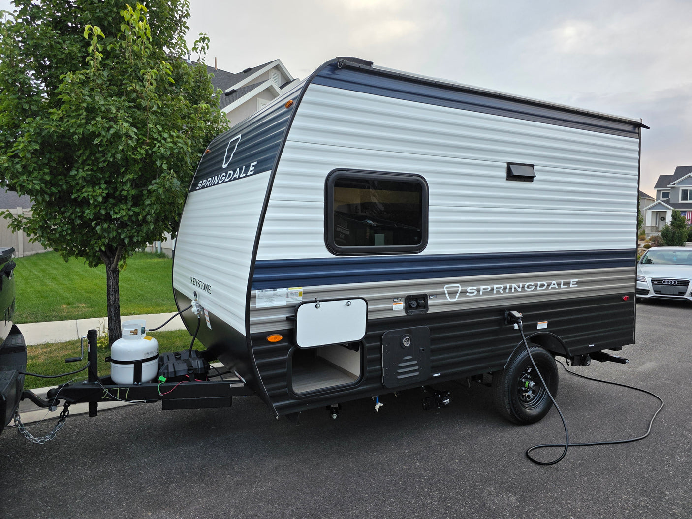 A white and blue Springdale travel trailer, ideal for your next rv rental, parked on asphalt with a tree and houses.