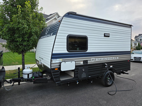 A white and blue Springdale travel trailer, ideal for your next rv rental, parked on asphalt with a tree and houses.