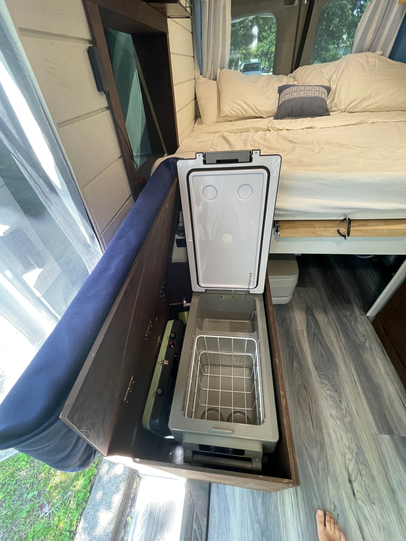 RV rental interior view: an open portable fridge/cooler, a small stove, and a neatly made bed.
