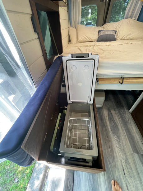 RV rental interior view: an open portable fridge/cooler, a small stove, and a neatly made bed.