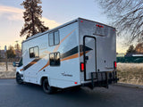 Side/rear view of a white/grey Coachmen Cross Trail RV, ideal for an rv rental, parked on asphalt with a rear cargo rack.