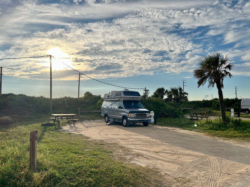 Silver RV rental camper van parked at a sandy campsite with a picnic table and palm tree under a bright, cloudy sky. — photo 11