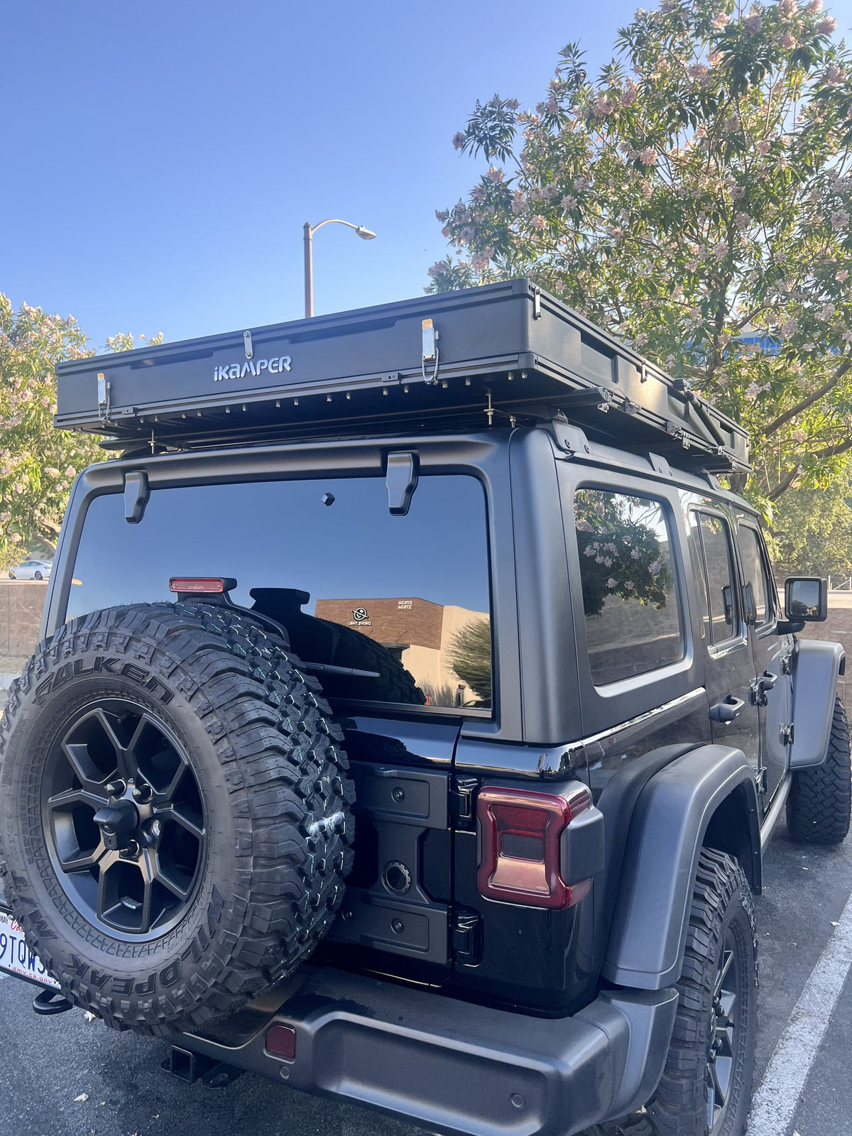 iKamper BDV Duo hardshell rooftop tent closed on its roof rack above a black Jeep Wrangler Unlimited, shot from the rear three-quarter in bright daylight with the iKamper logo visible along the shell side — photo 1