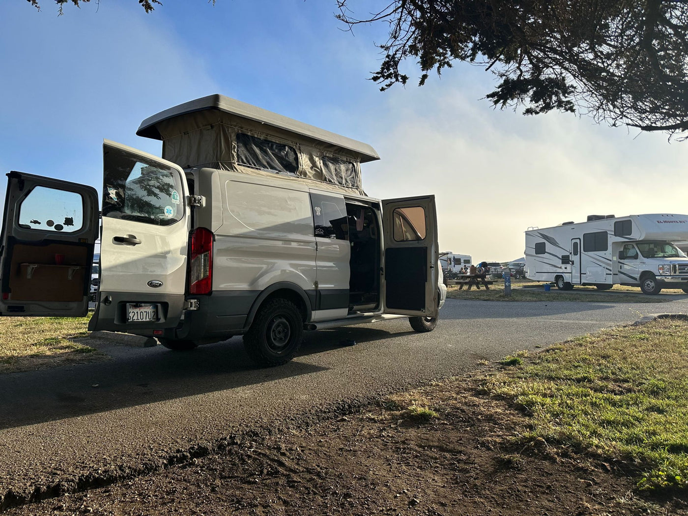  parked by the ocean at sunset. An RV rental offering stunning coastal views for your adventure.|A white pop-top camper van is parked with its doors open — photo 2