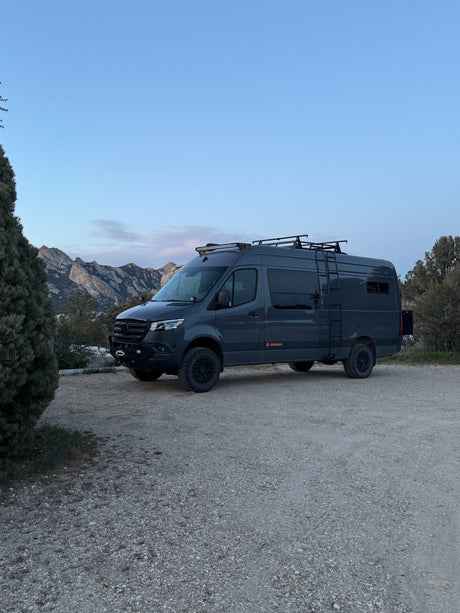 Adventure-ready Mercedes Sprinter camper van for rv rental, parked on gravel with mountains & trees in the background.