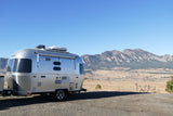 An Airstream RV rental is parked on gravel, facing majestic mountains and a sprawling plain under a bright blue sky.
