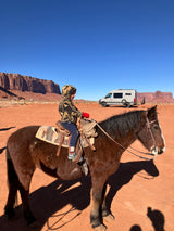 A child on horseback, wearing red gloves, in a vast desert landscape with red rock formations. A light gray RV rental is parked nearby.